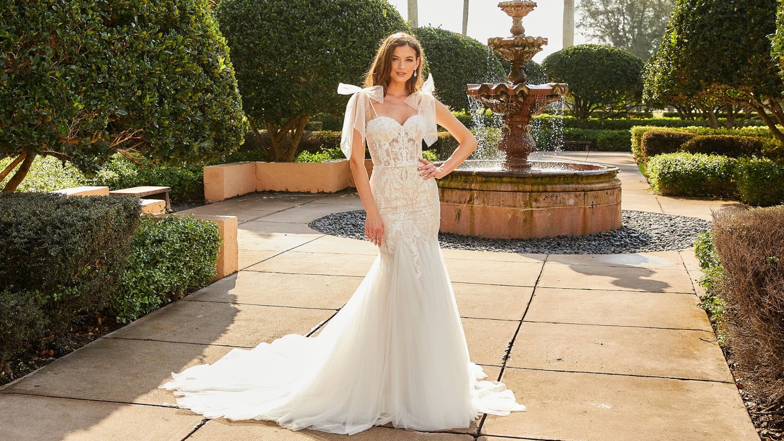 Bride in an elegant lace wedding gown stands on a sunlit patio with neatly trimmed greenery and a flowing fountain, conveying a serene and joyful ambiance.