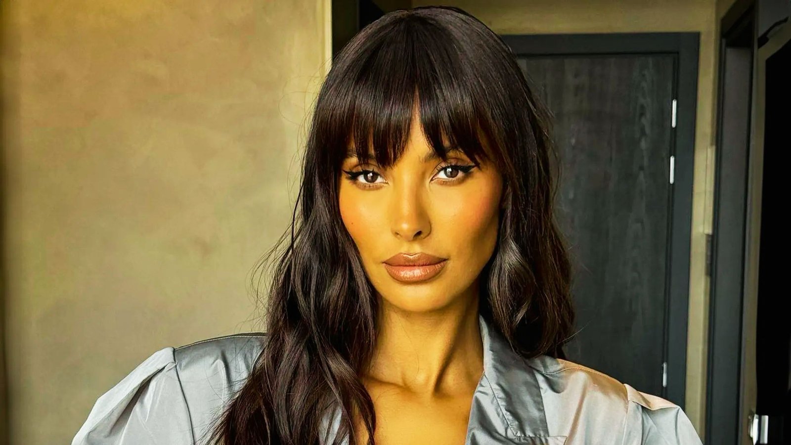 A woman with long layered hair and Bardot bangs, wearing a silver shirt, poses confidently.
