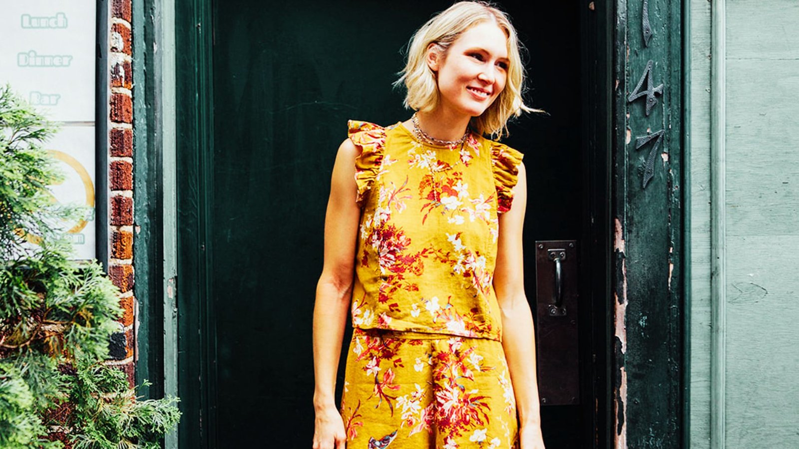 A woman in a yellow floral dress stands confidently in front of a door, showcasing a stylish matching set outfit.