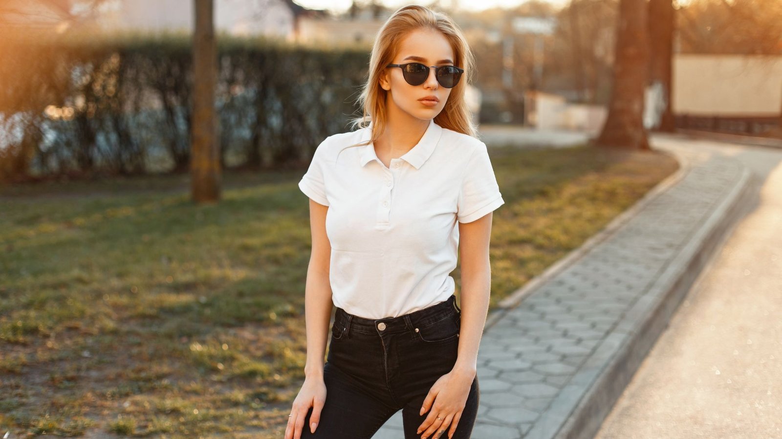 Woman in a chic effortless mom outfit wearing a crisp white polo shirt tucked into high-waisted black skinny jeans, accessorized with black sunglasses on a sunny sidewalk.