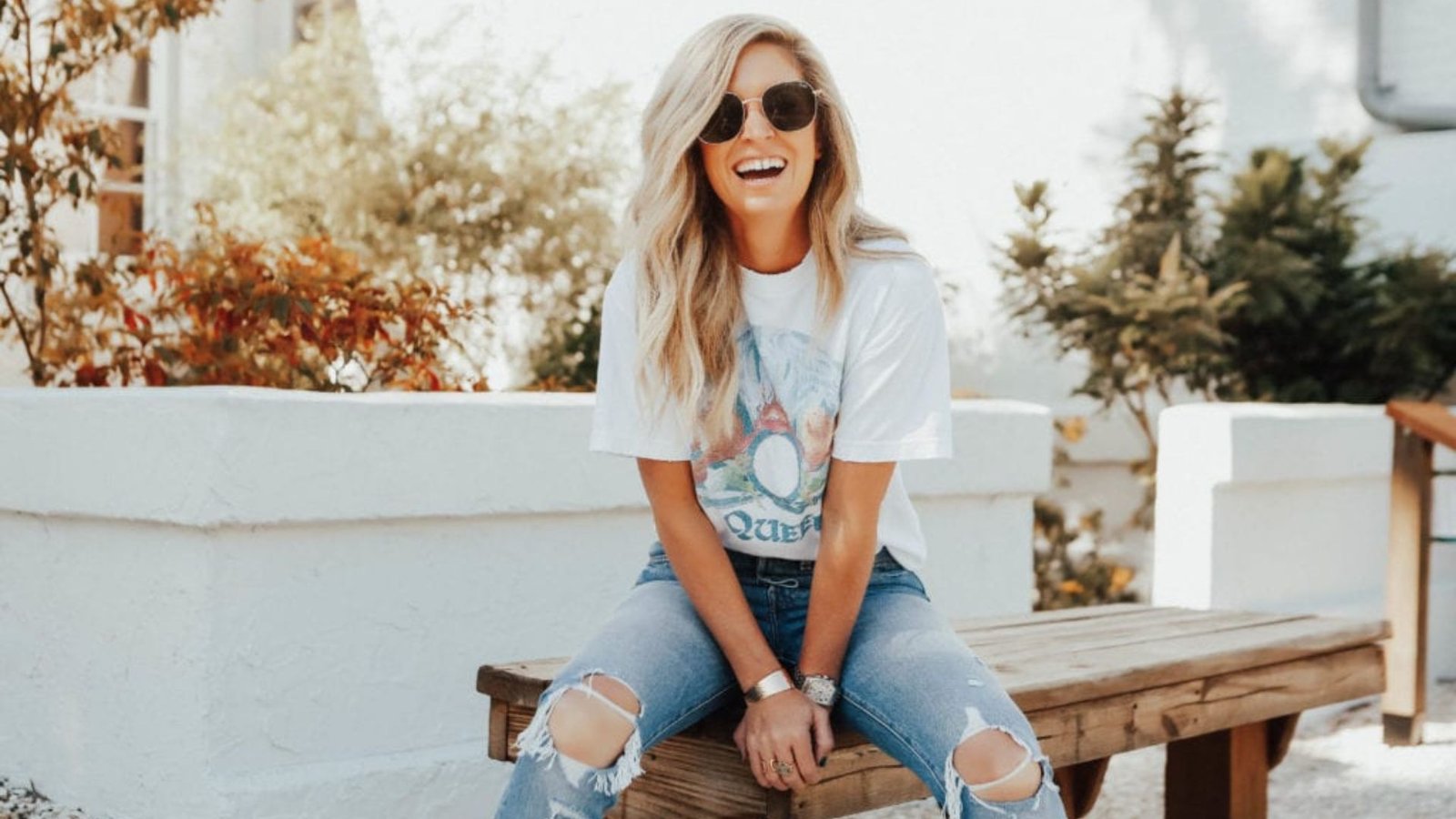 Smiling woman in a white graphic tee and ripped light-wash jeans sitting on a bench.