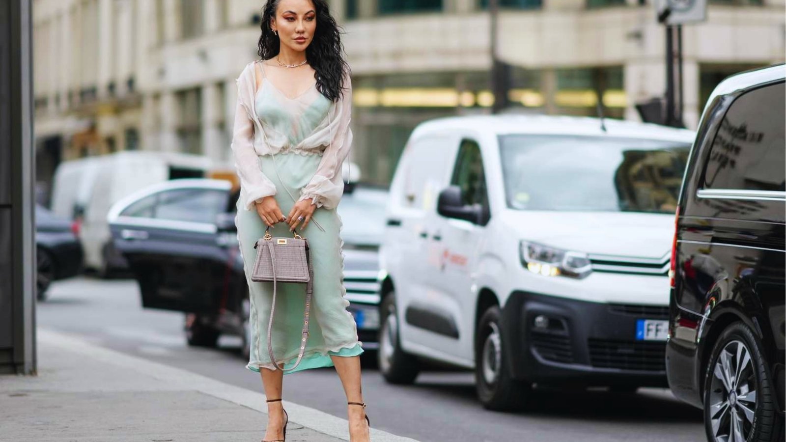 A woman stands confidently on a city street in a sheer green dress, holding a small handbag. Cars and vans are visible in the background, creating an urban atmosphere.