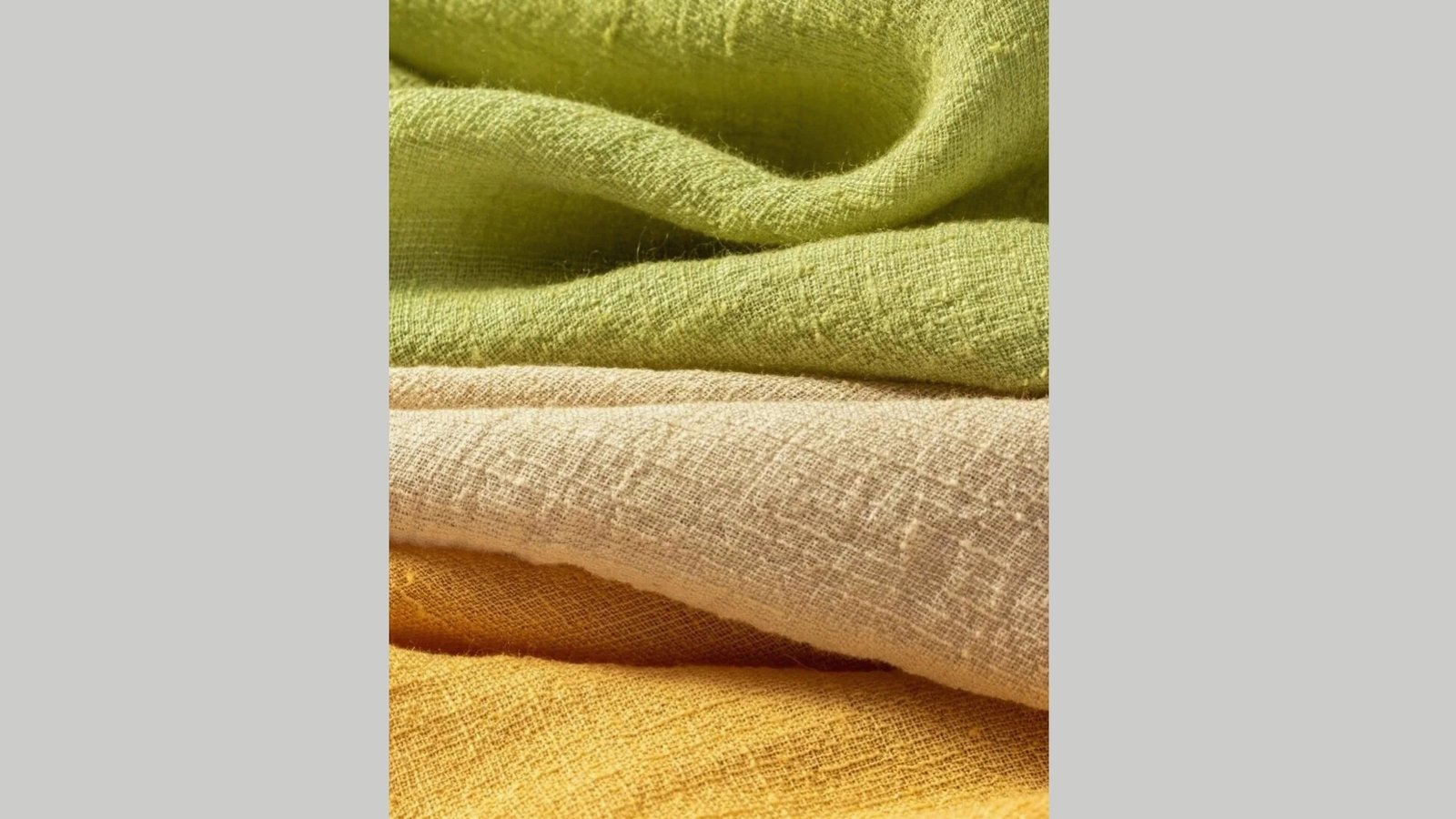 Close-up of layered linen fabrics in green, beige, and yellow. The textures are visibly coarse, conveying warmth and softness. Natural and cozy tone.