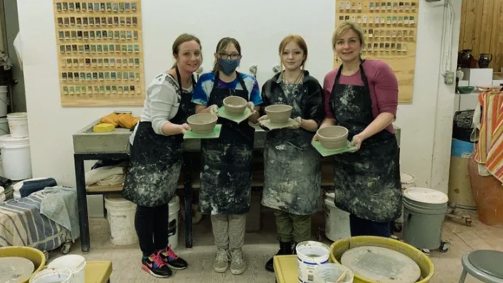 Four people in pottery class proudly display their clay bowls. They wear aprons and stand in a workshop setting, exuding creativity and teamwork.