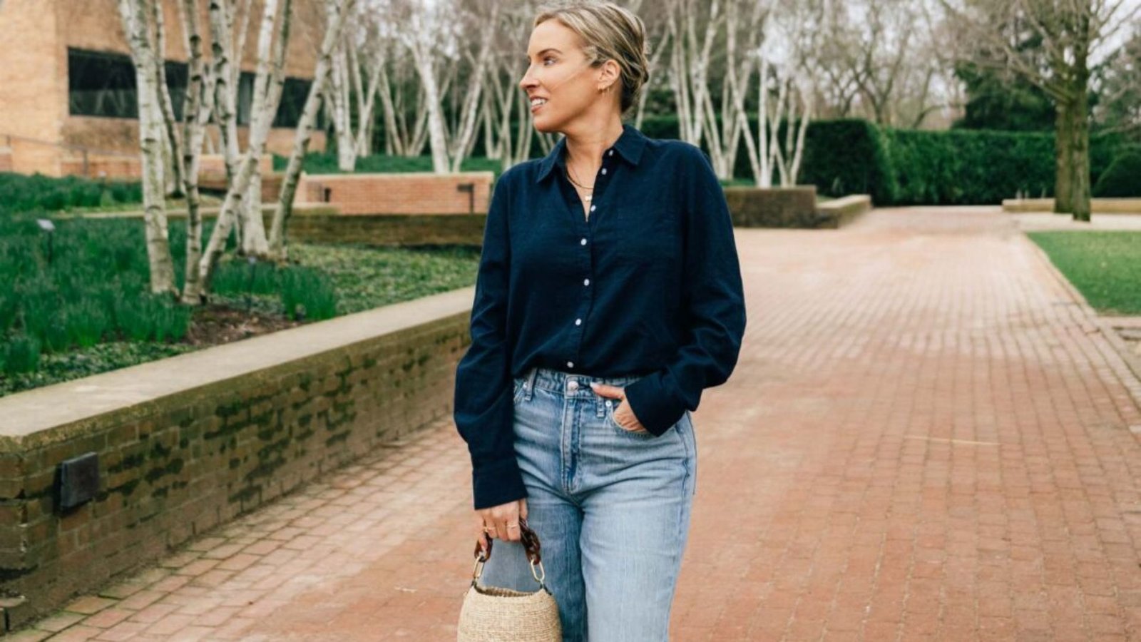 Smiling woman wearing a navy blue button-down shirt tucked into light wash straight-leg jeans, holding a woven straw bag while walking on a brick pathway for an effortless mom outfit.