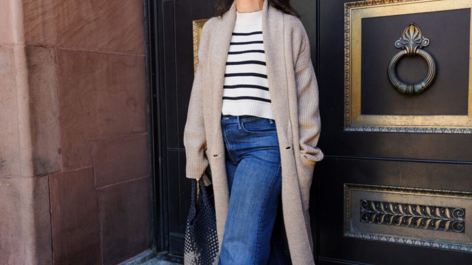 Woman in beige longline cardigan layered over a black-and-white striped sweater and blue jeans. A classic 90s mom outfit blending comfort and structure for casual days.