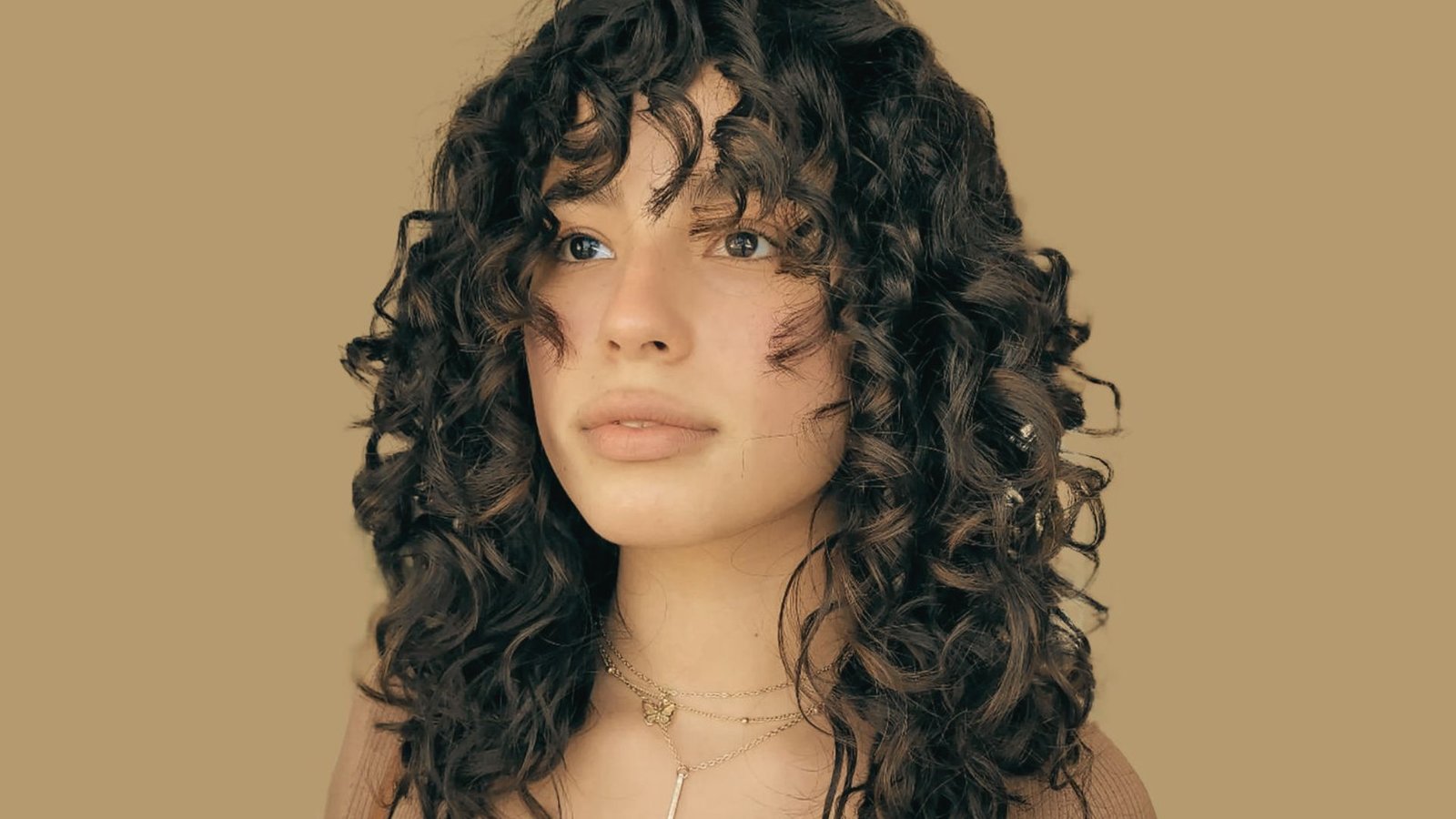 A woman with natural curly hair and bangs, wearing a stylish necklace, smiles at the camera.