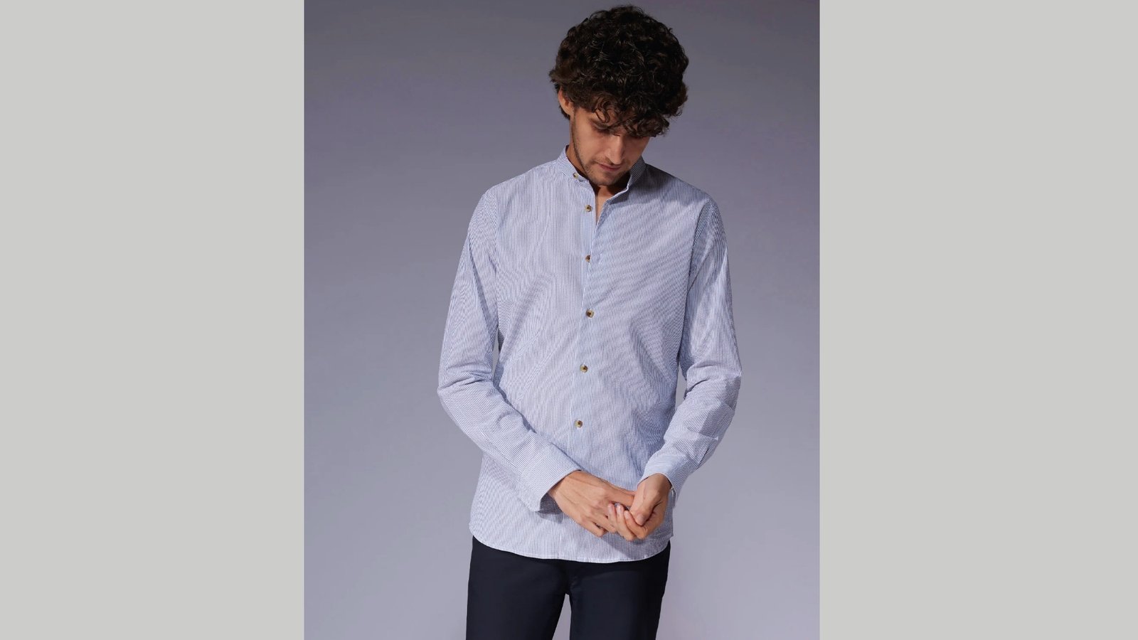 A man with curly hair wears a light blue, striped button-down shirt and dark pants, adjusting his sleeve. The background is plain gray, conveying a calm tone.