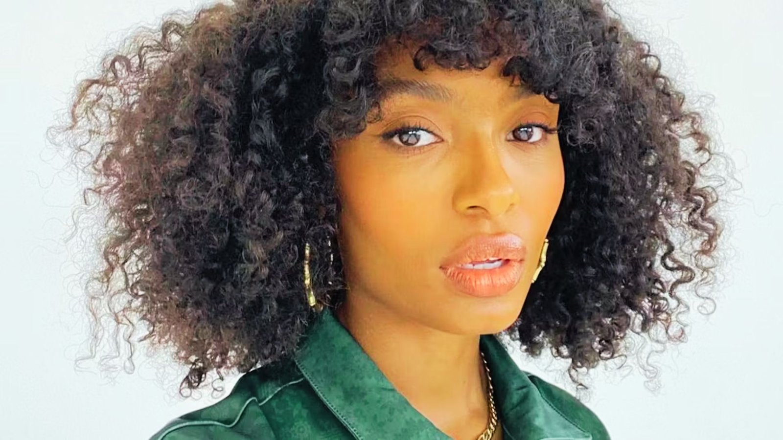 A woman with curly hair wearing a green shirt, showcasing Bardot bangs styled for curly hair.
