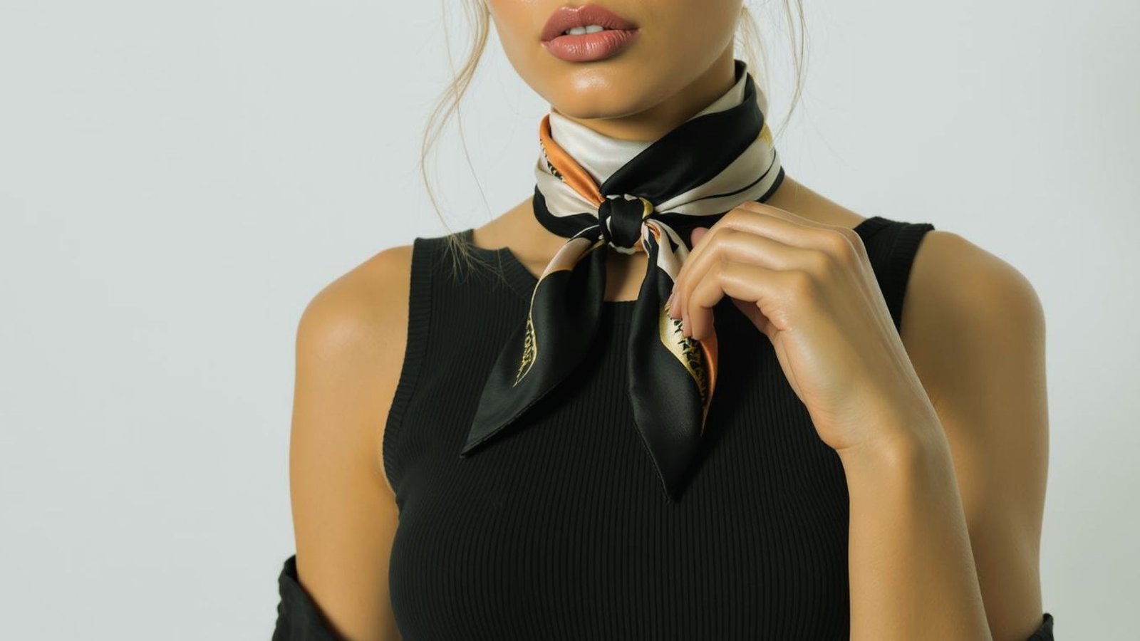 A woman in a black sleeveless top wears a black and beige silk scarf tied around her neck. She holds the scarf with one hand, exuding elegance and confidence.