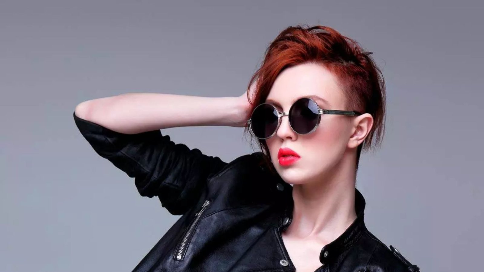 Woman wearing sunglasses and leather jacket with auburn hair styled up. A short undercut on the side is visible, creating a modern punk-inspired look.