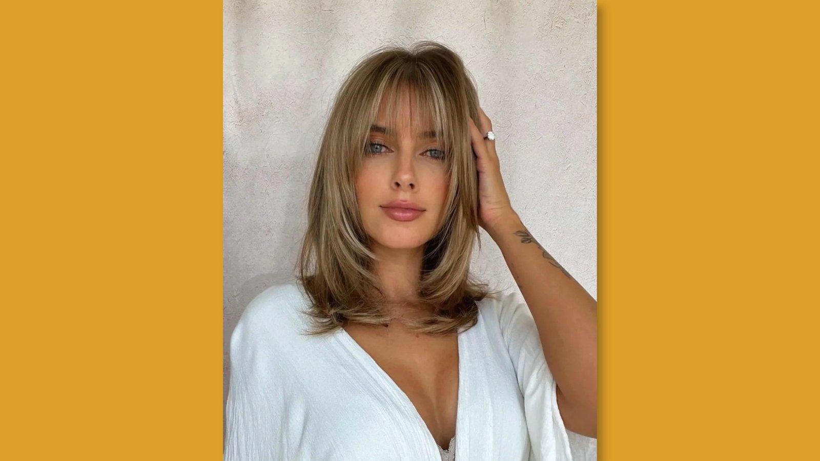 Woman with honey blonde shoulder length haircut featuring wispy curtain bangs and face-framing layers in white V-neck top