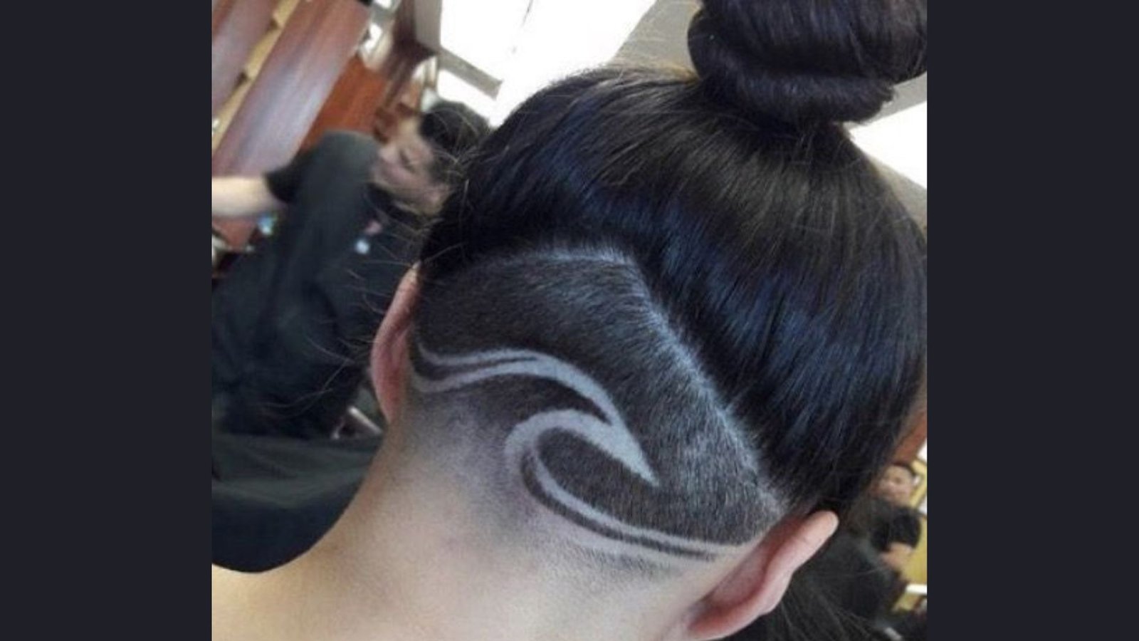 Back view of a woman with dark hair in a topknot revealing a wave design shaved into the nape undercut.