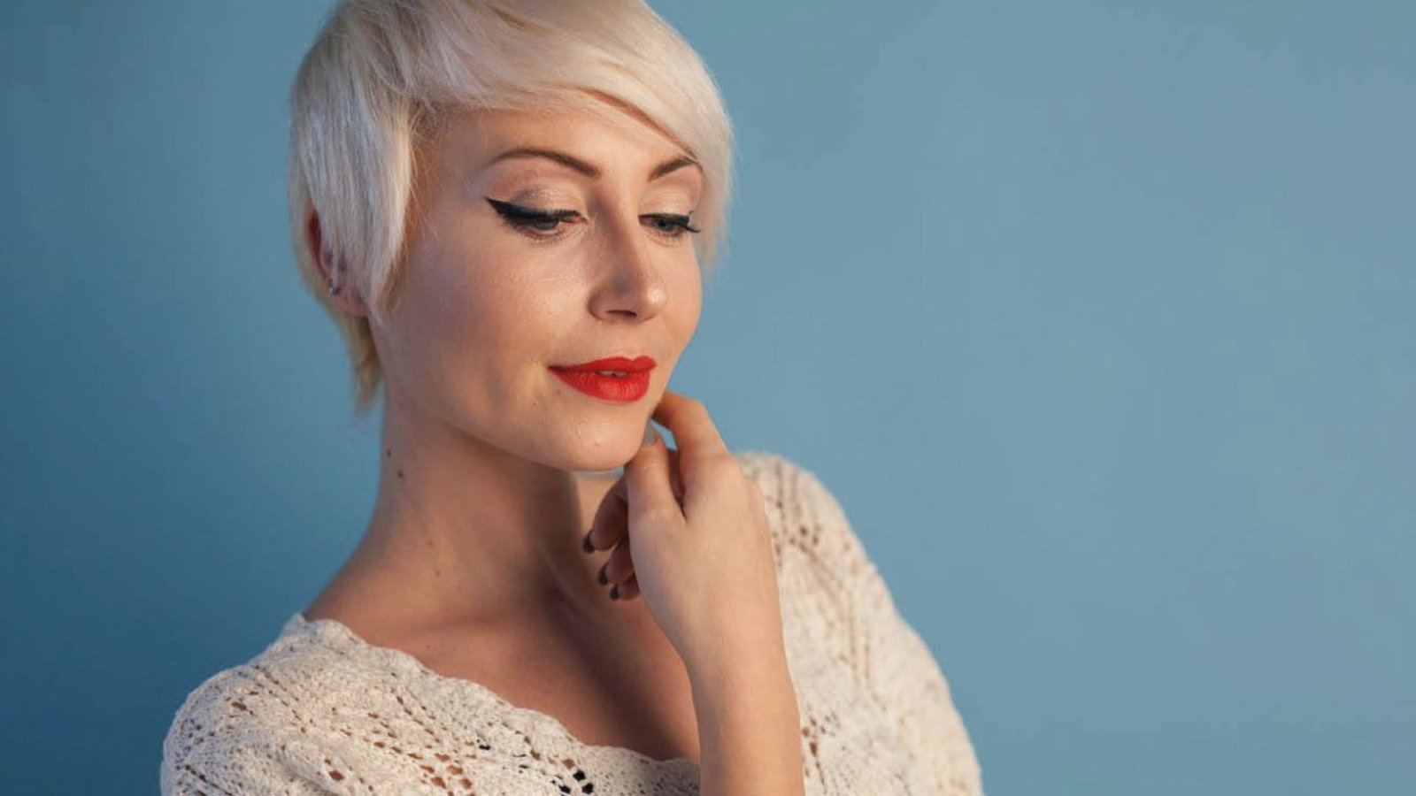 A woman with short blonde hair styled in a sleek yet textured pixie cut, wearing bold red lipstick.