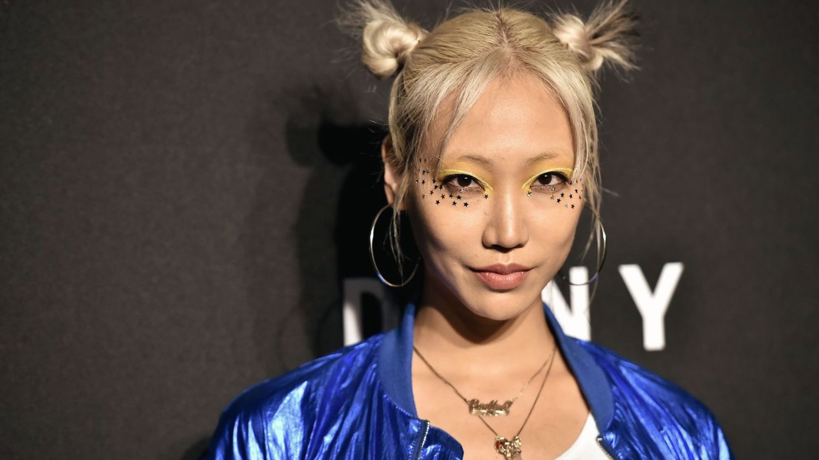A woman with blonde mini space buns and bangs, wearing a blue jacket.