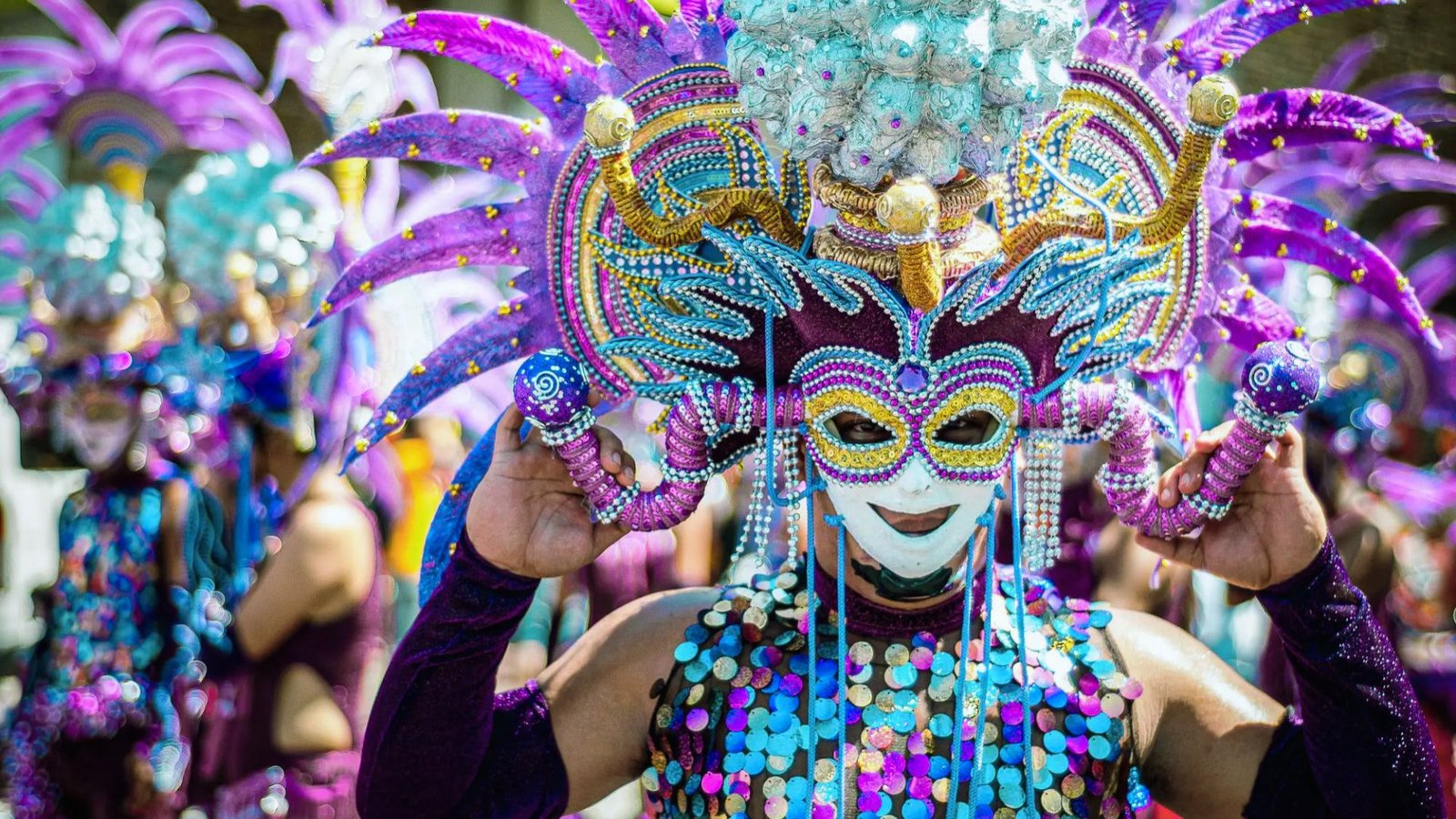 A vibrant festival scene with a person in an elaborate, colorful costume featuring a bejeweled mask and headdress, surrounded by others in similar attire.