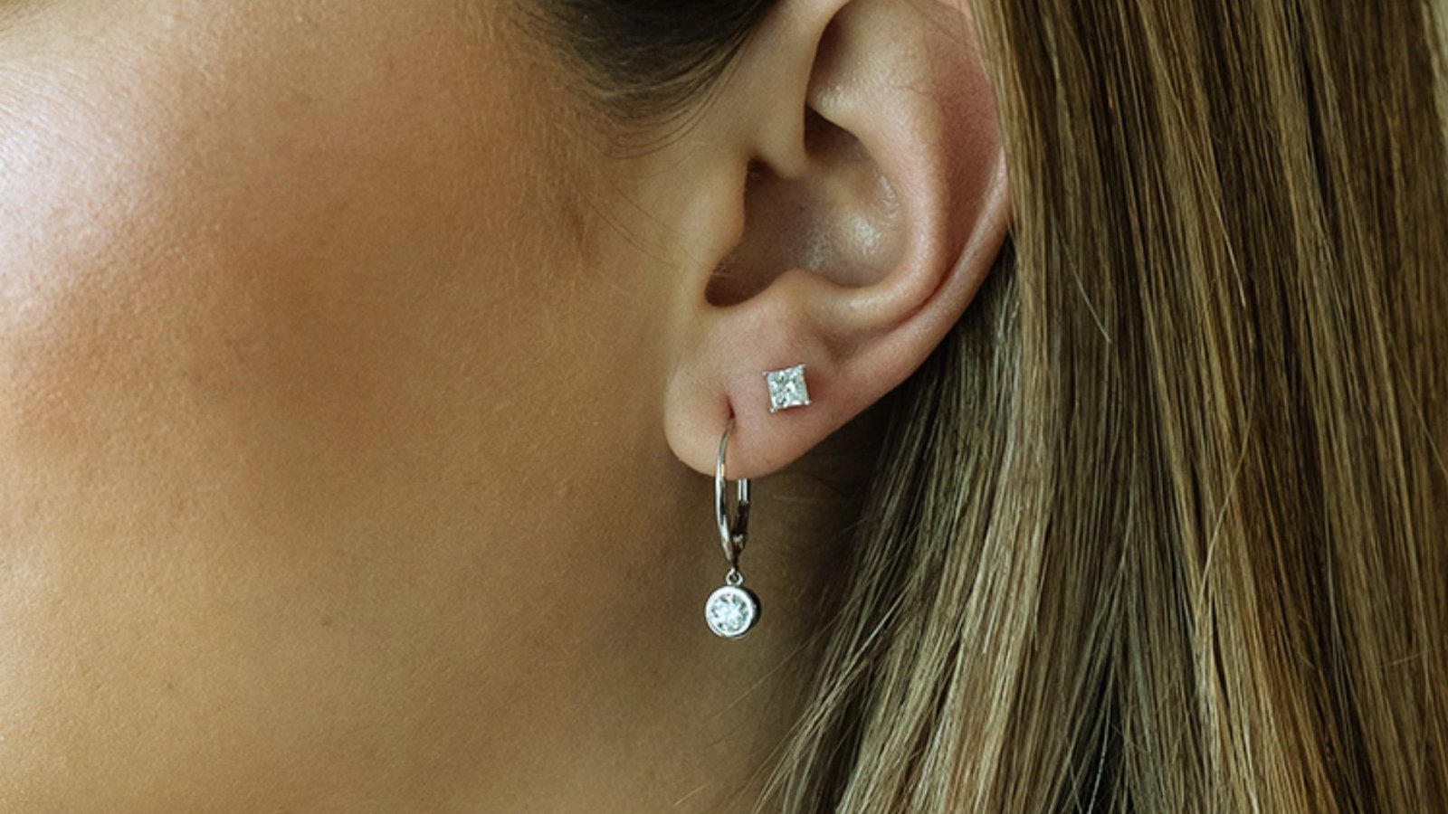 Close-up of an ear adorned with a small square stud earring and a dangling hoop earring with a crystal. Blonde hair partially visible to the right.