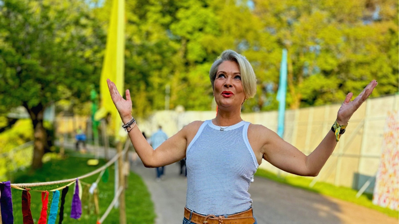 Woman with short blonde hair raises her arms joyfully in a park. She's wearing a striped tank top, surrounded by trees and colorful flags.