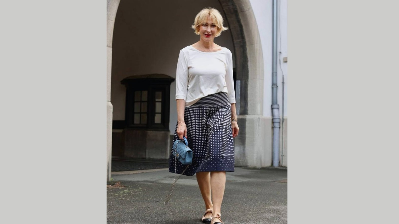 A woman with short blonde hair walks confidently on a pavement. She wears a white shirt and a blue patterned skirt, holding a blue handbag. The background features an archway with a classical architectural style, adding elegance to the scene.