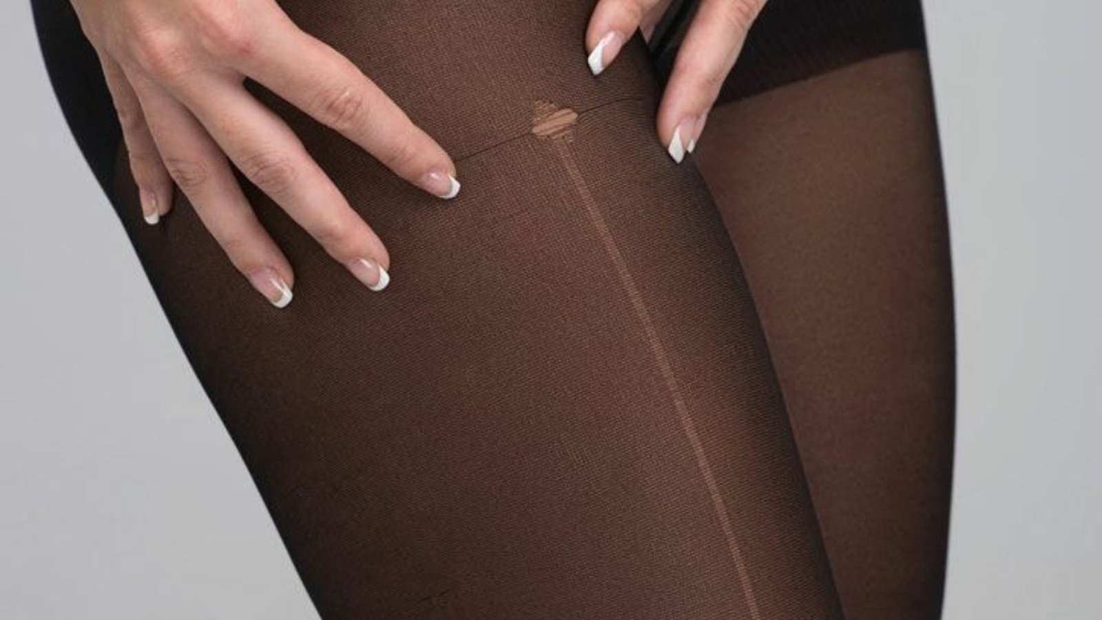 Close-up of hands with French manicured nails touching a run in sheer black pantyhose. The image conveys a sense of frustration or inconvenience.