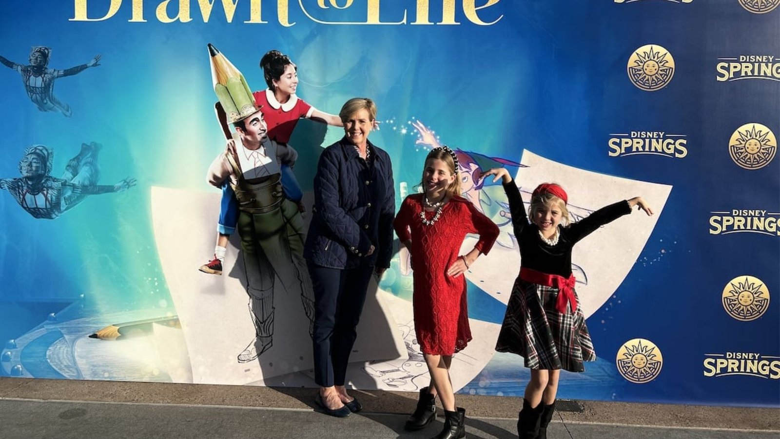 A woman and two children pose joyfully in front of the "Drawn to Life" backdrop at Disney Springs. The background features vibrant blue hues, whimsical characters, and dynamic elements, conveying excitement and creativity.