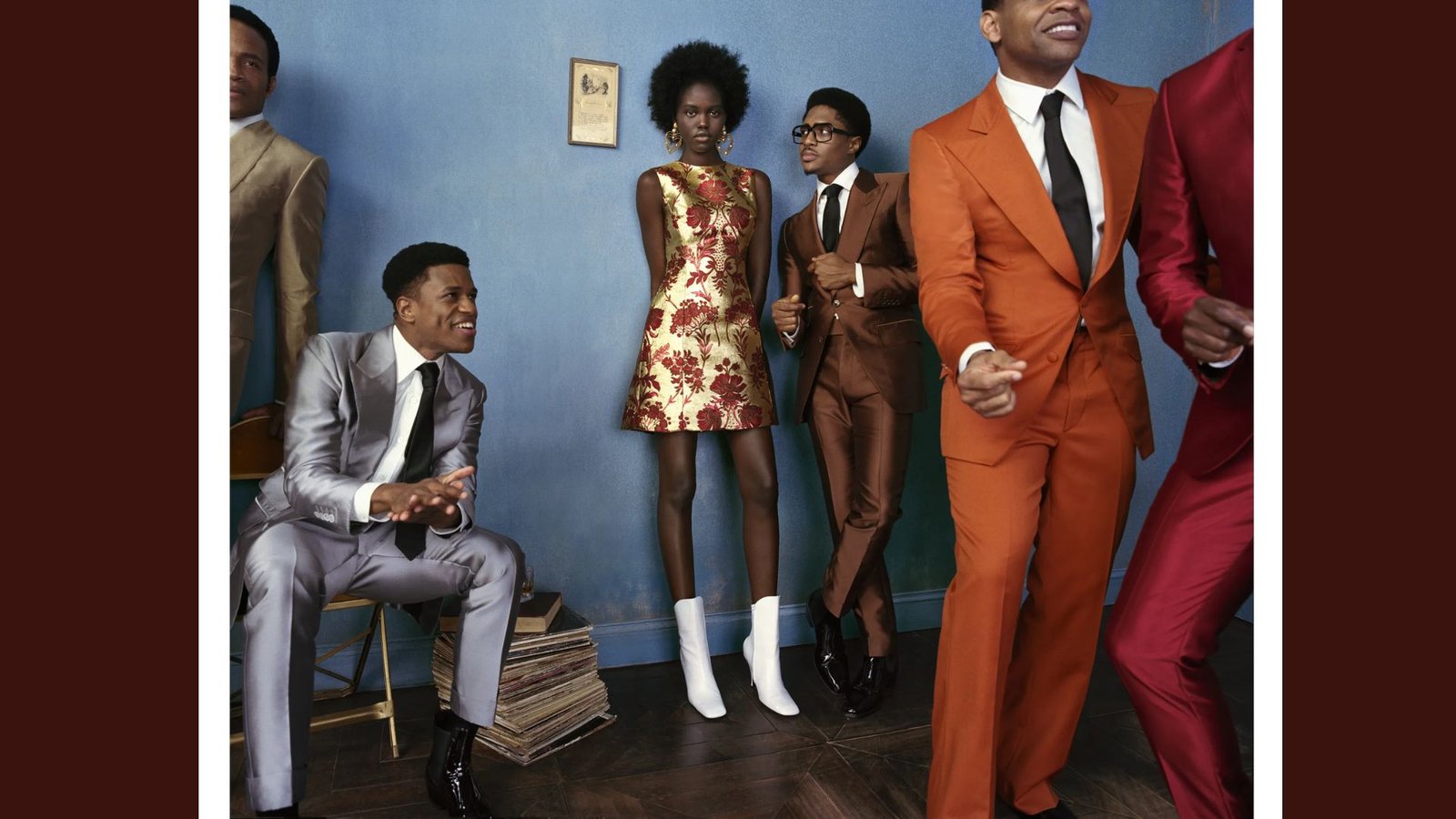 A stylish group from the 1960s stands against a blue wall, wearing vibrant suits and a floral dress. Their lively expressions suggest joy and celebration.