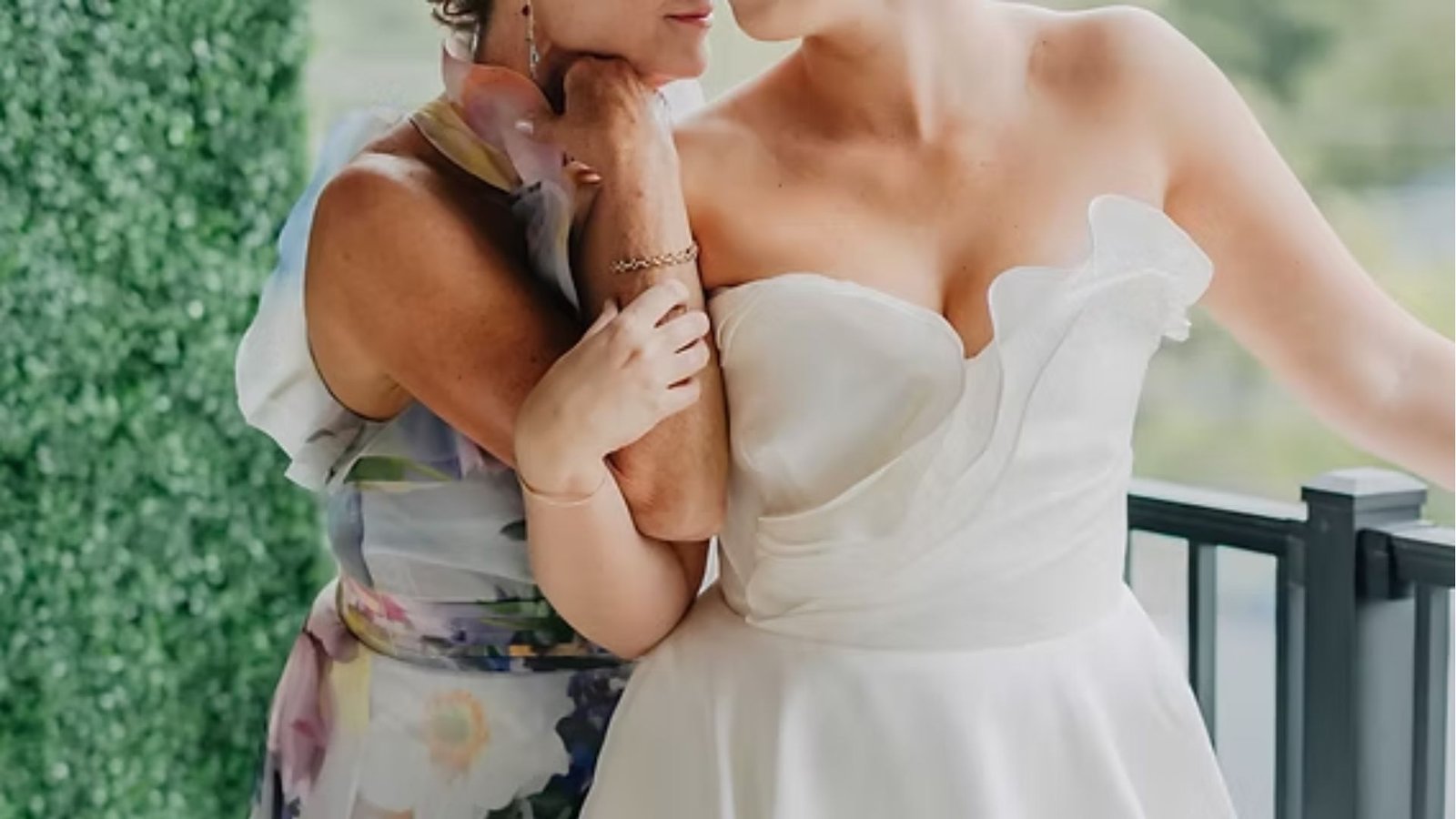 Bride in a strapless white dress embraces a woman in a floral gown, conveying warmth and affection. They stand on a balcony with greenery.