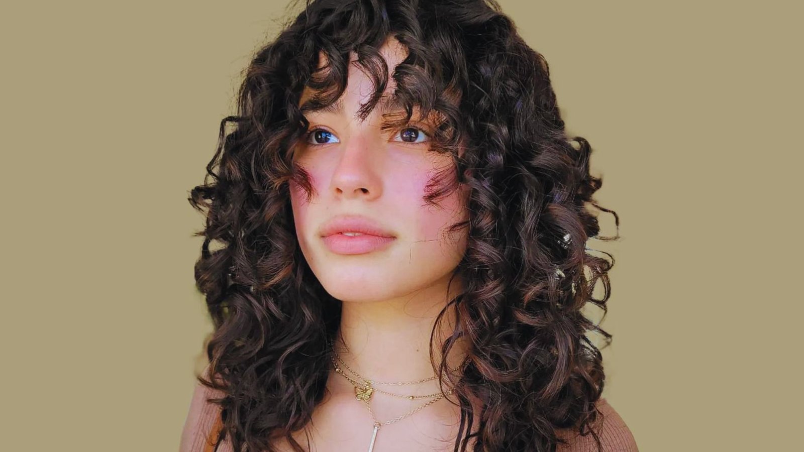 A woman with curly hair and curtain bangs, wearing a stylish necklace, poses for a hair styling tips guide.