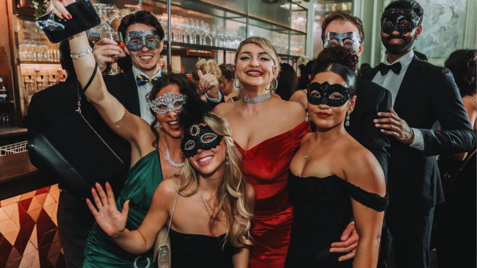 A group at a masquerade party poses joyfully for a selfie. They wear elegant masks and formal attire, exuding a festive and lively atmosphere in a stylish bar setting.