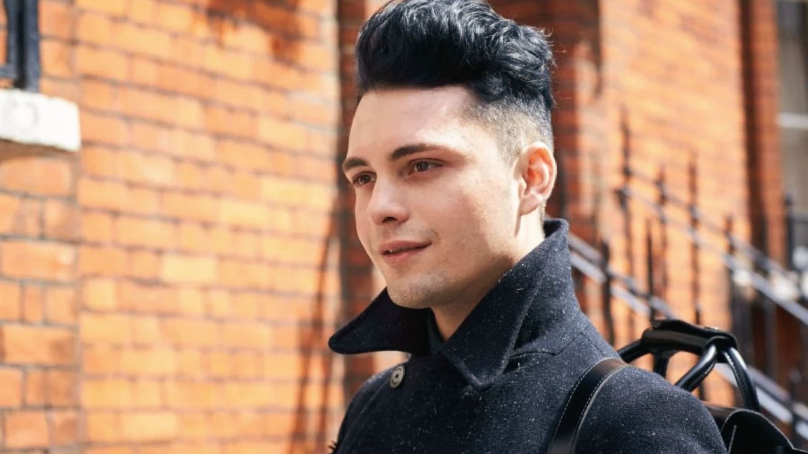 A man wearing a black jacket and backpack, sporting a messy disconnected undercut fade hairstyle.