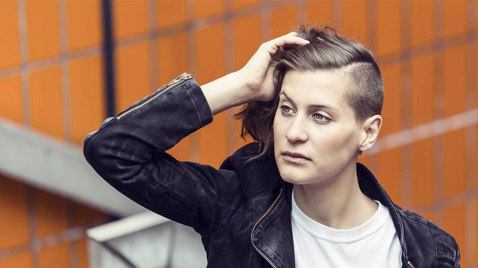 Woman in black leather jacket running hand through short brown hair with shaved side undercut. The longer top is swept over to create contrast against the buzzed side panel.