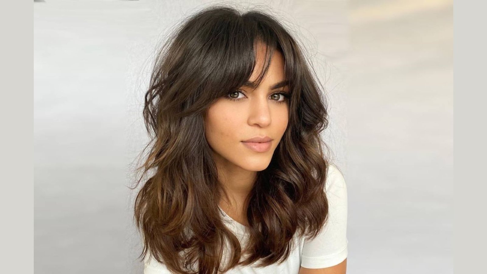 Woman with shoulder-length wavy brunette hair and thick curtain bangs. The fringe parts softly in the center and blends into face-framing layers.