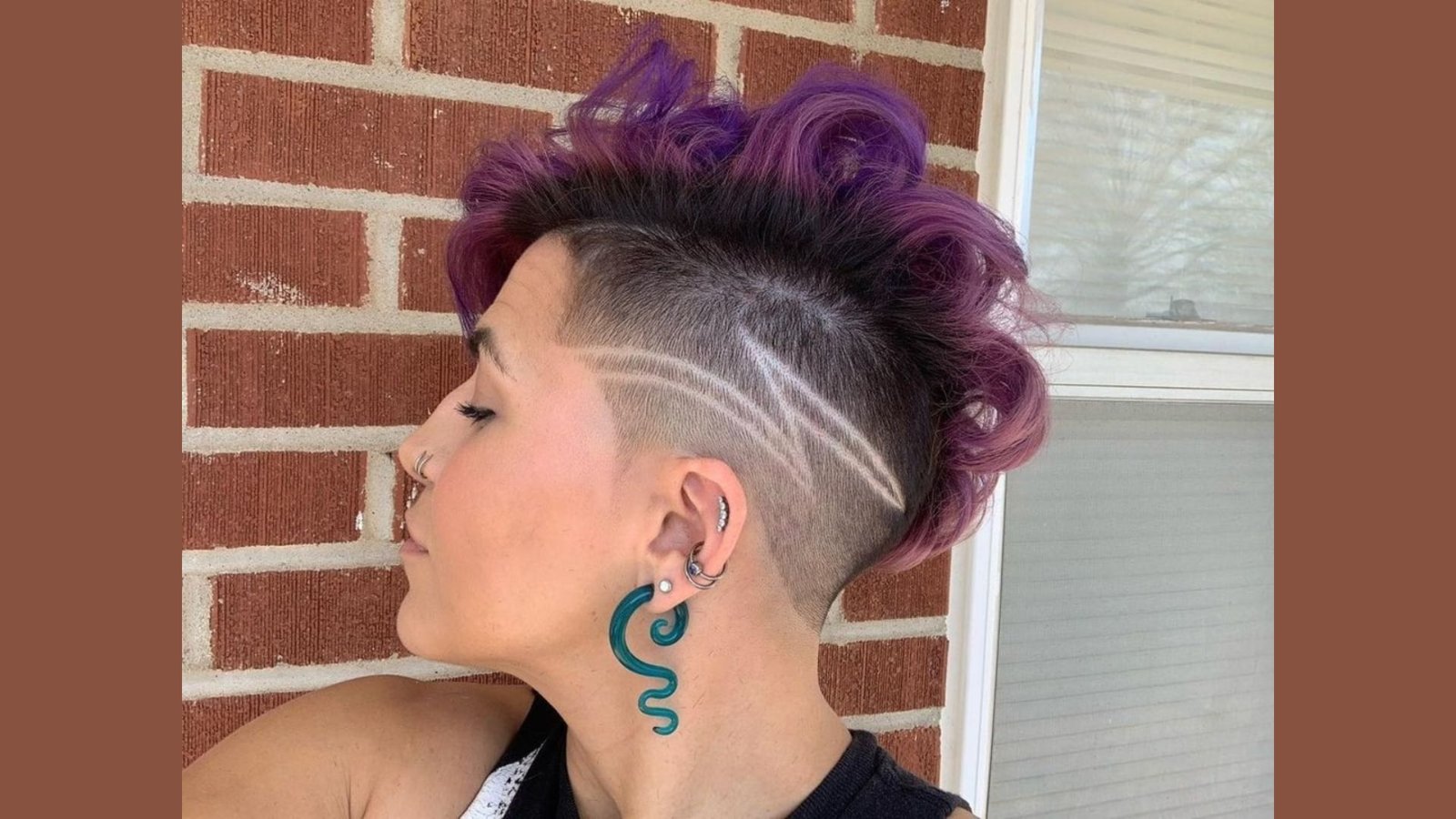Profile of a woman with a curly purple faux hawk and shaved side undercut with line designs.