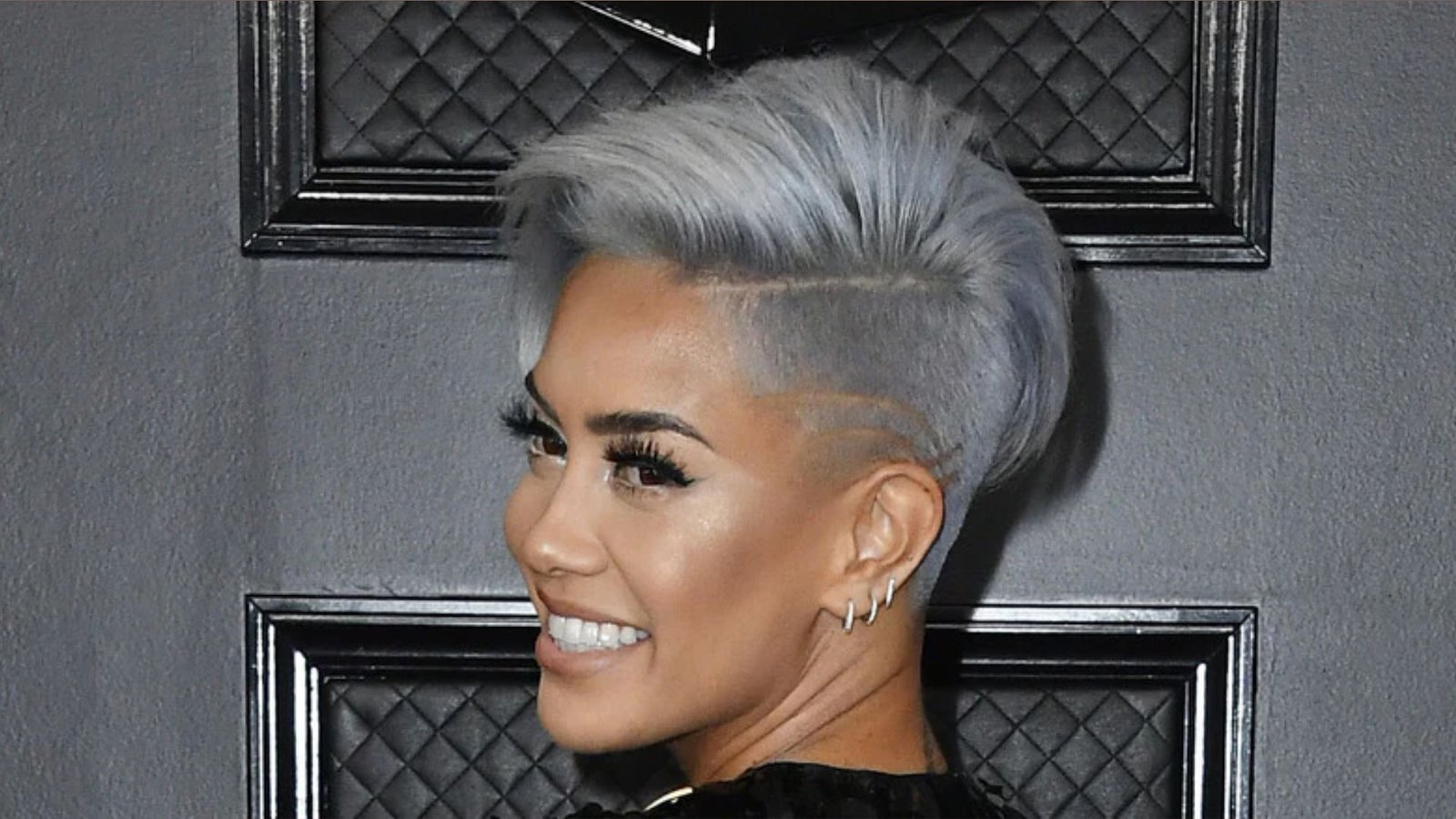 Smiling woman at a formal event showing a silver-gray undercut pixie with shaved side designs. A voluminous swept top paired with closely cropped sides creates a bold red-carpet hairstyle.