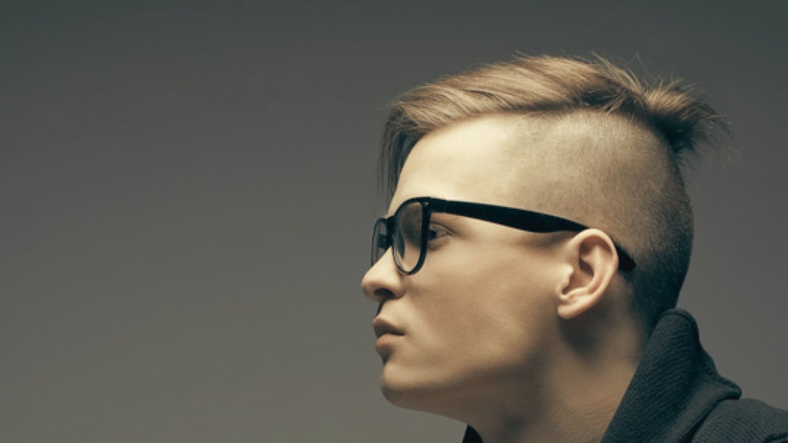A man with glasses and a mohawk hairstyle gazes thoughtfully to the side, showcasing a half-up disconnected undercut fade.