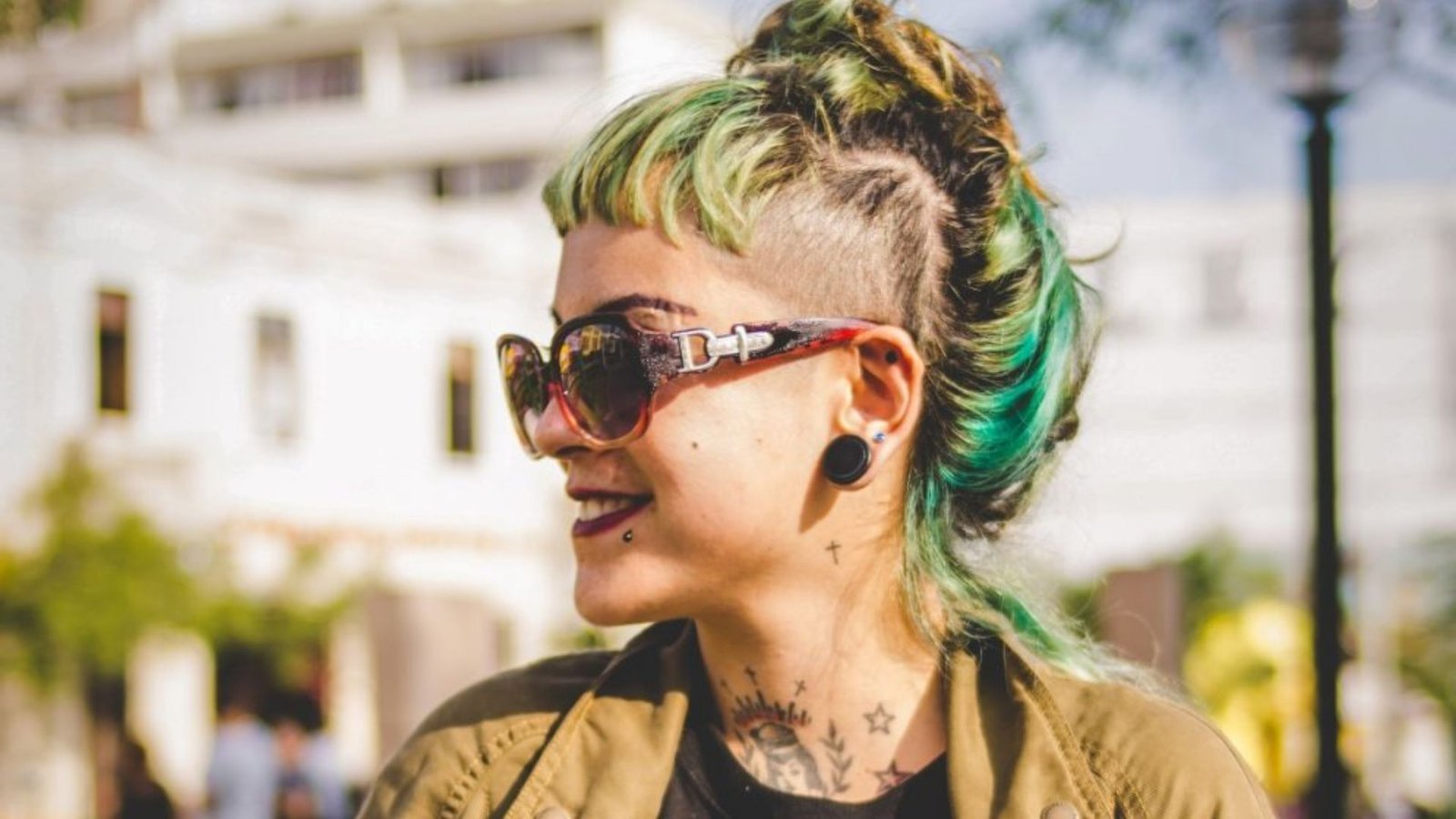 Side view of a person with green-black hair in a topknot and shaved side undercut. Bold micro bangs and visible neck tattoos pair with the disconnected buzzed temple.