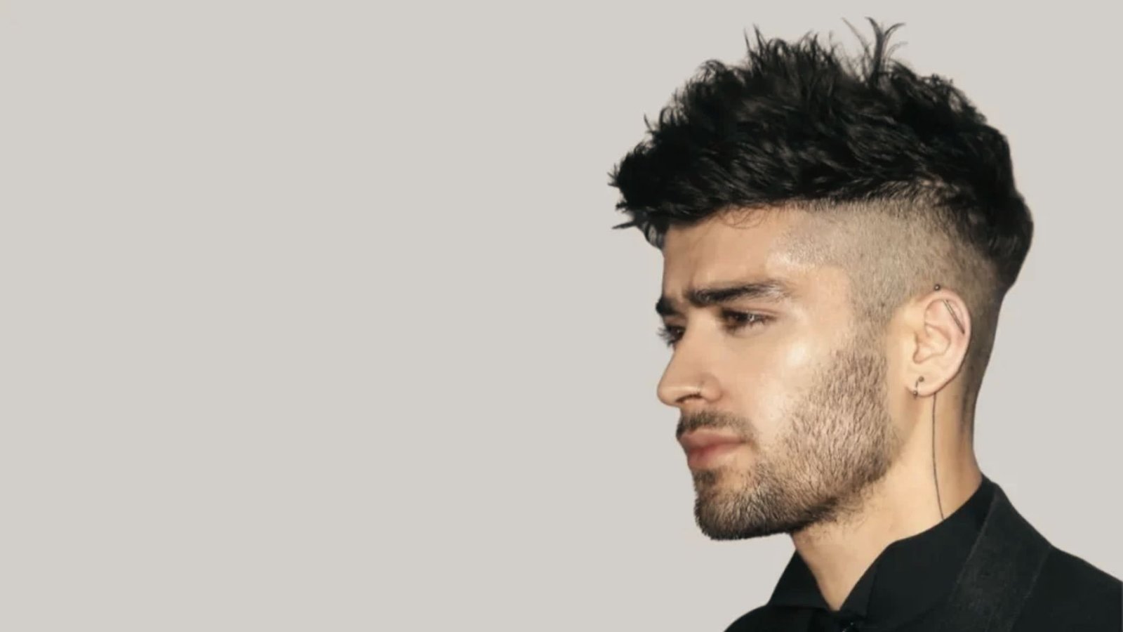 A man in a black suit with a short spiky disconnected undercut fade, looking away from the camera.