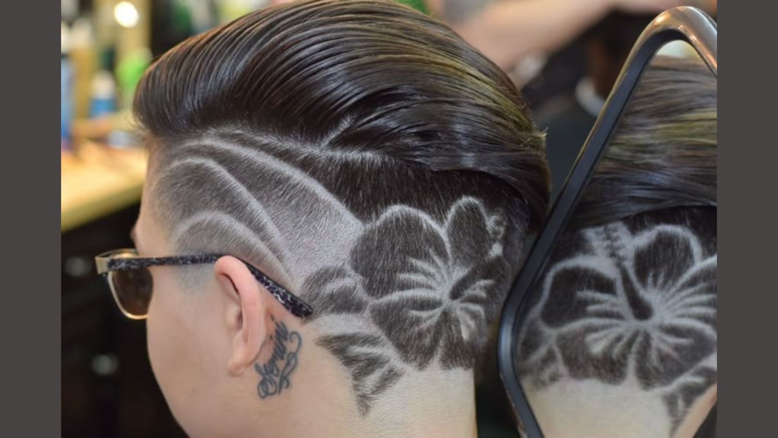 Side view of a person with slicked-back dark hair and elaborate hibiscus flower design shaved into the side undercut. Detailed floral artwork is visible in the mirror reflection, paired with a neck tattoo and glasses.