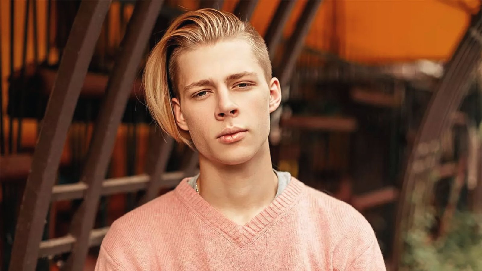 A young man with blonde hair styled in a side-swept fringe, wearing a pink sweater.