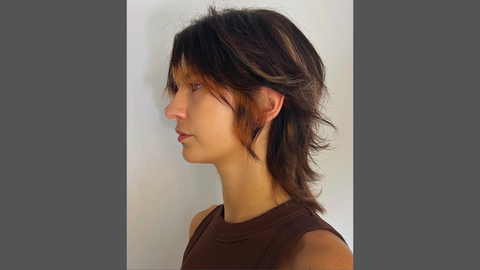 Side profile of woman with midi wolf-cut lite haircut featuring choppy layers, wispy bangs, face-framing highlights, and textured length in brown hair.