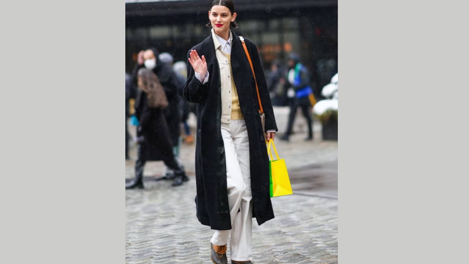 A woman in a long black coat waves confidently while walking on a rainy city street. She wears white pants, a cream sweater, and carries a bright yellow bag.