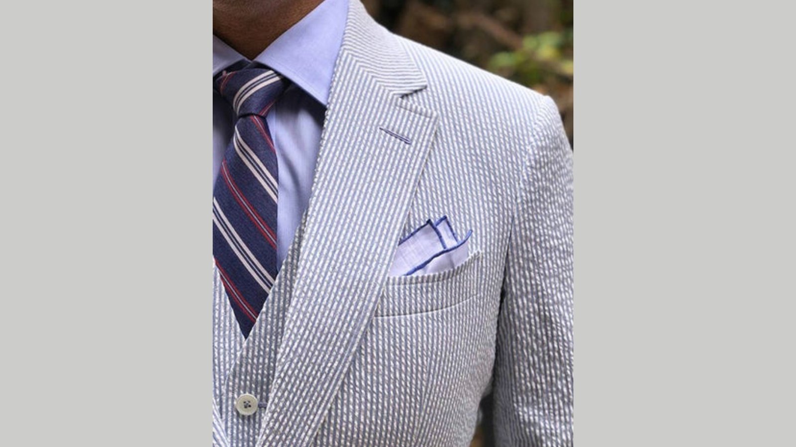 Man in a light blue pinstripe suit with a lavender shirt and striped tie; a folded pocket square peeks from the breast pocket, conveying elegance.