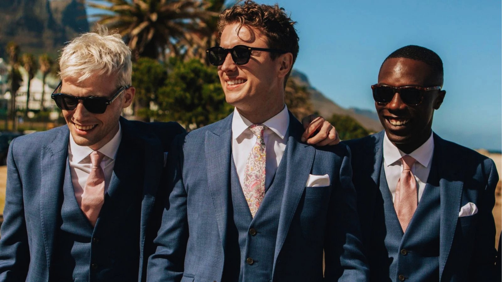 Three men in blue suits with pink ties and sunglasses smile joyfully against a backdrop of palm trees and a clear blue sky, exuding a cheerful vibe.