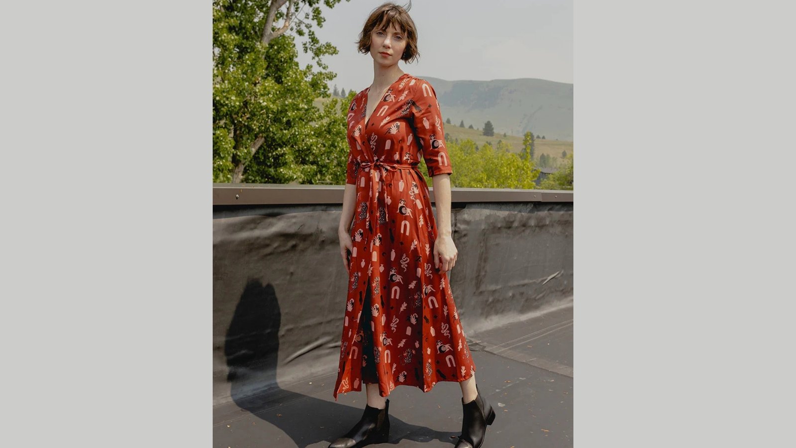 Woman in a red patterned dress stands on a rooftop. The dress has long sleeves and a belt. She wears black boots. Green trees and hills are in the background.