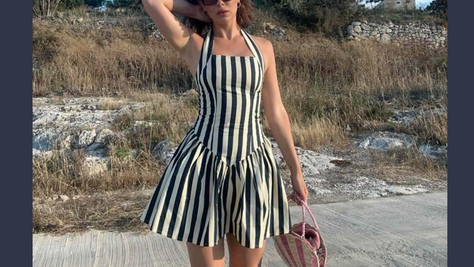 A woman stands confidently on a path, wearing a black and white striped dress. She holds a pink woven bag, with dry grass and rocky terrain in the background.