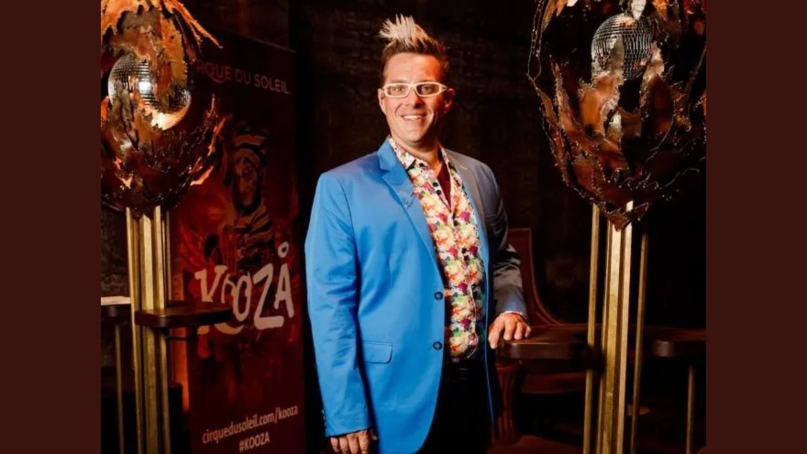 A person wearing a bright blue blazer over a floral shirt smiles confidently. They stand between two intricate, artistic sculptures. The backdrop features a colorful "Kooza" poster from Cirque du Soleil, evoking a vibrant, theatrical atmosphere.