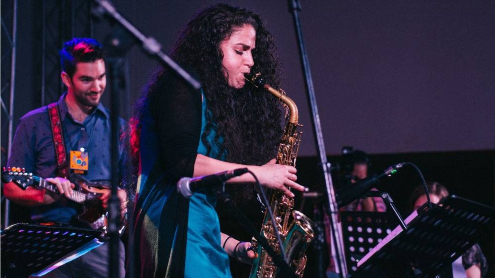 A woman intensely plays the saxophone on stage with a guitarist in the background. The setting is lively, evoking a sense of passion and enthusiasm for music.