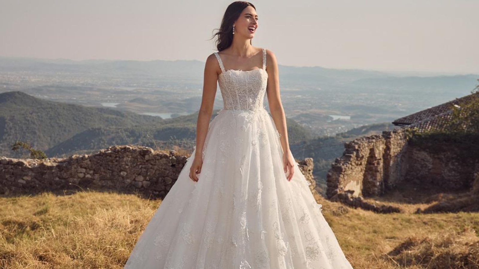 A woman in a flowing white wedding dress stands smiling in a grassy field with mountains and ancient stone ruins in the background, exuding joy and serenity.
