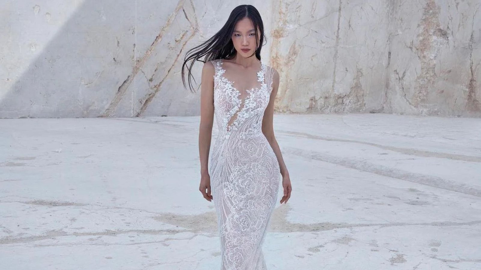 A woman in a lace gown walks on a white marble background. Her long hair flows as she gazes forward. The scene is elegant and serene.