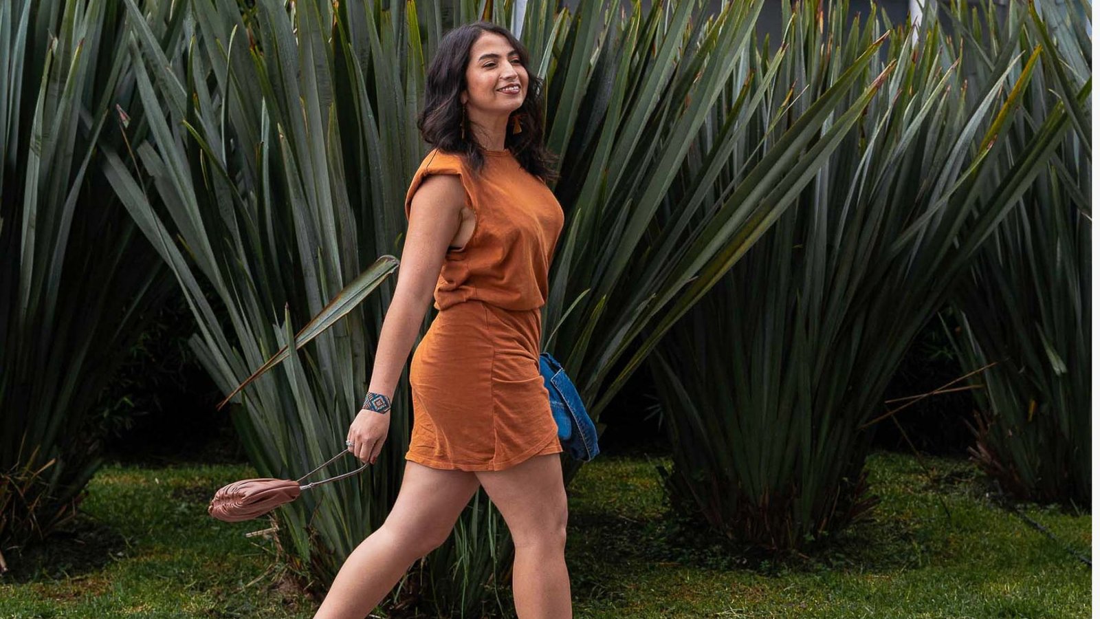 Smiling woman walking outdoors in a trendy rust orange sleeveless mini dress with padded shoulders, holding a small brown clutch and denim jacket for an effortless mom style outfit.